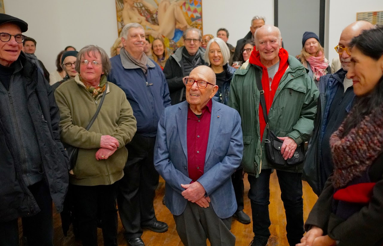 Painter Philip Pearlstein standing at the center of an art gallery looking off to the side surrounded by people. 